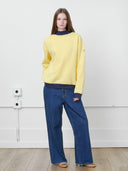 Female model wearing ONE DNA butter yellow mockneck sweatshirt with navy blue contrast ribbed cuffs and hem, paired with relaxed blue jeans.